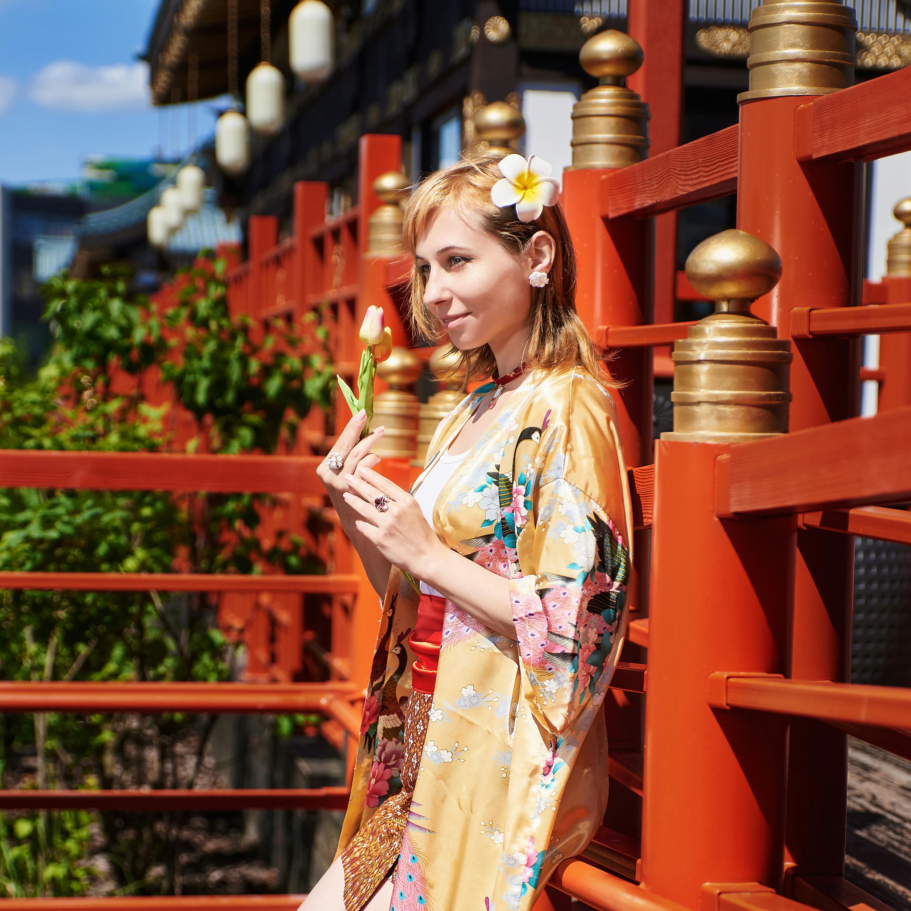 Vrouw in gele kimono met bloem in haar haar, houdt een tulp vast op een traditionele Japanse brug. Zonnige dag, rode houten reling, serene sfeer.