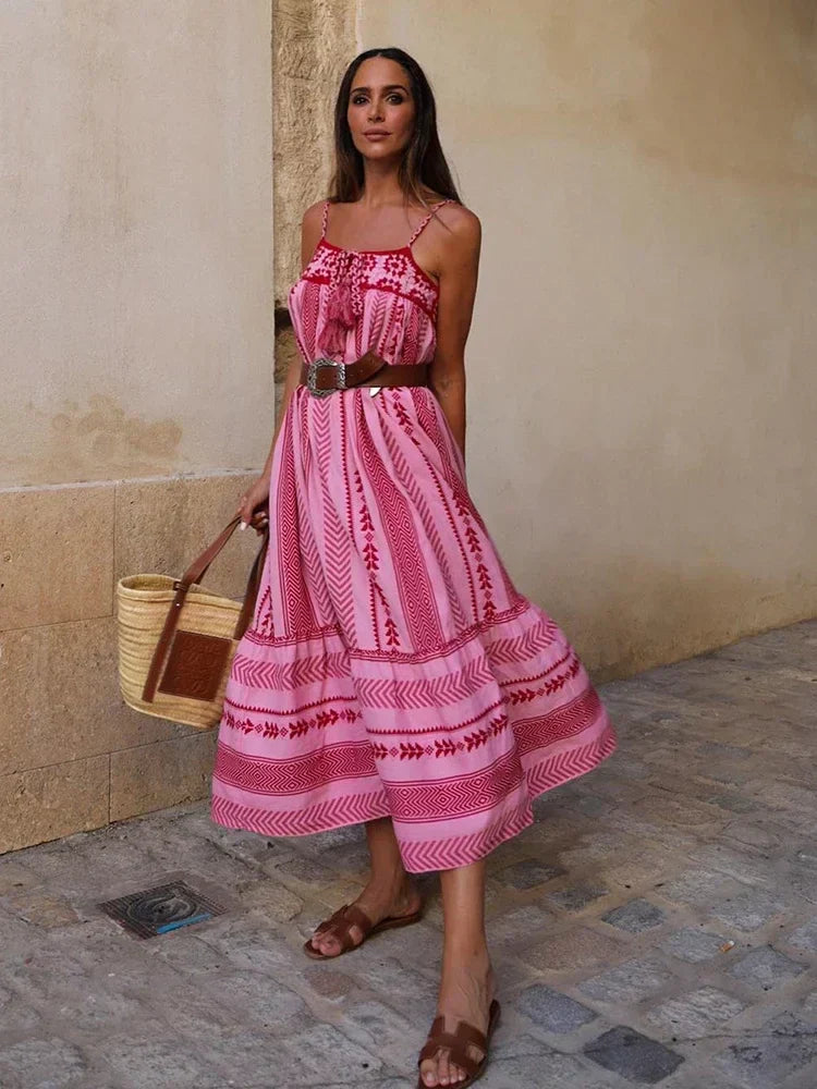 Vrouw in roze bohemian zomerjurk met spaghettibandjes, bruine riem en sandalen, draagt een rieten tas. Perfect voor zomerse mode en boho-stijl.