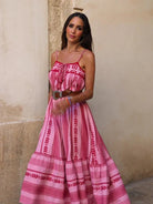 Vrouw in roze bohemian maxi-jurk met spaghettibandjes, staand tegen een beige muur. Zomerse mode, boho-stijl, casual elegantie.