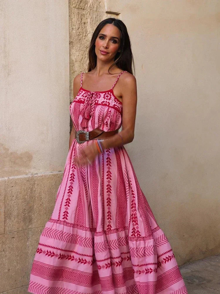 Vrouw in roze bohemian maxi-jurk met spaghettibandjes, staand tegen een beige muur. Zomerse mode, boho-stijl, casual elegantie.