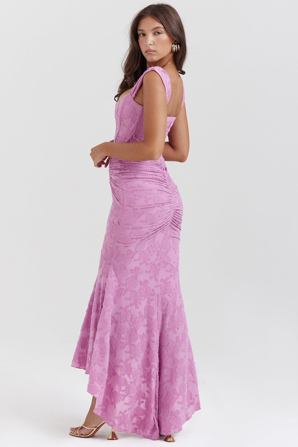 Vrouw in elegante roze kanten avondjurk met ruches, staand zijwaarts. Perfect voor galafeest, bruiloft of formele gelegenheid. Stijlvolle mode.