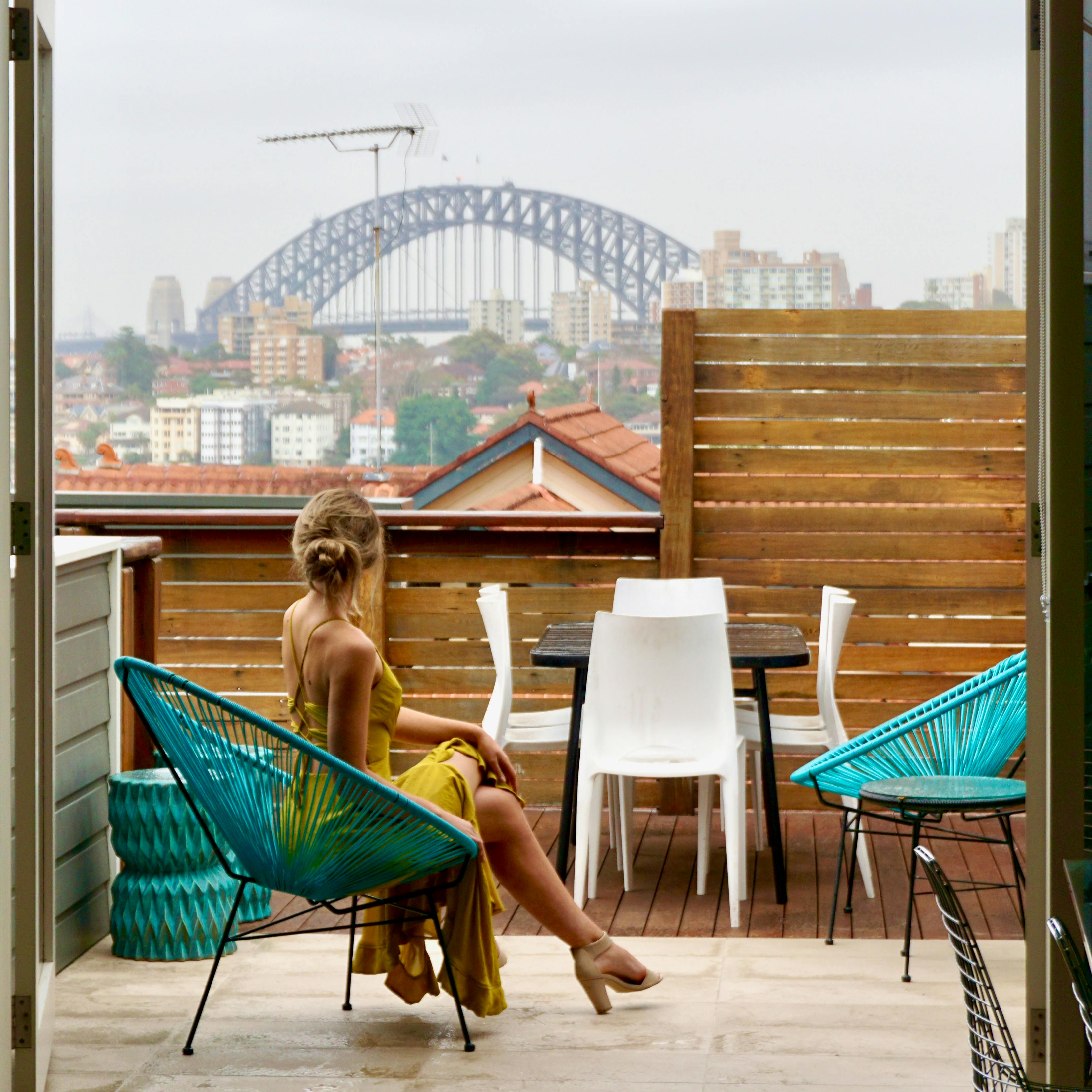 Vrouw in gele jurk op balkon met uitzicht op Sydney Harbour Bridge, moderne buitenmeubels, stedelijk landschap, ontspannen sfeer.